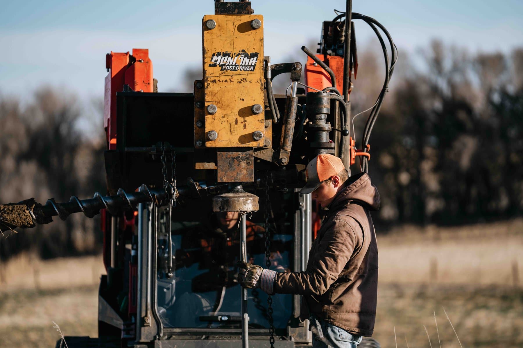 Operator running a hydraulic post driver mounted on equipment in open pasture.