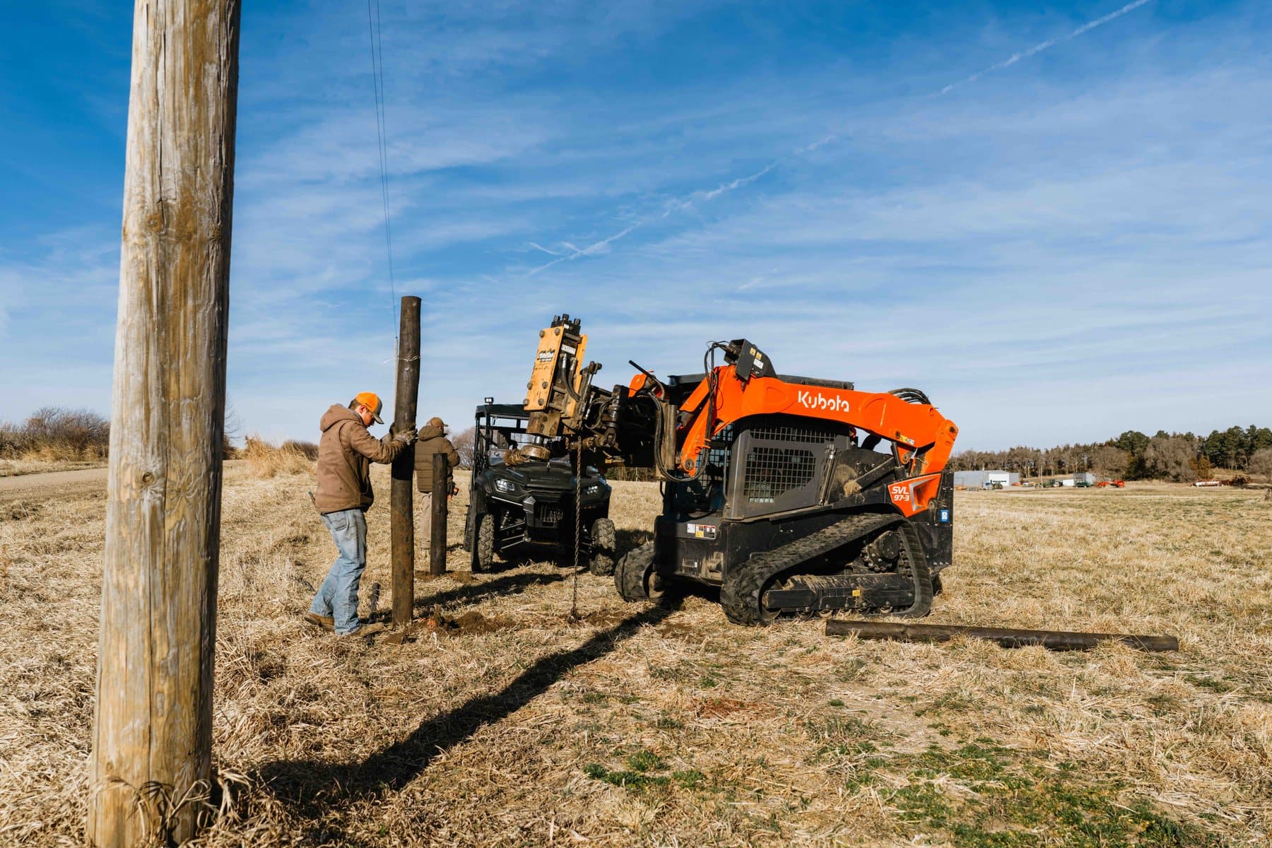 Crew setting posts with a skid steer while running barbed wire in open pasture.